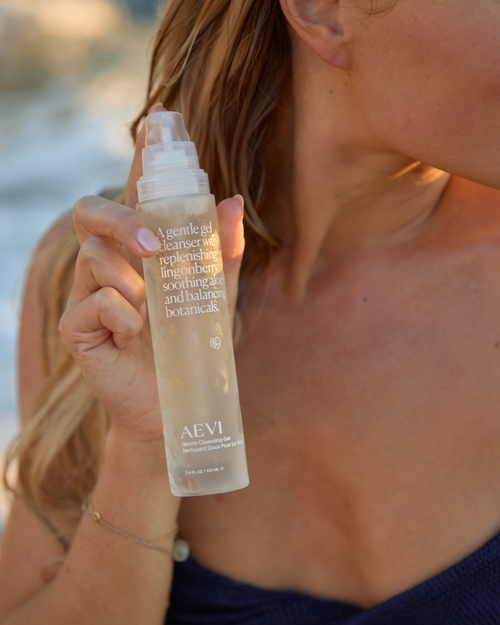 Woman holding a bottle of Aevi skincare product by the ocean.